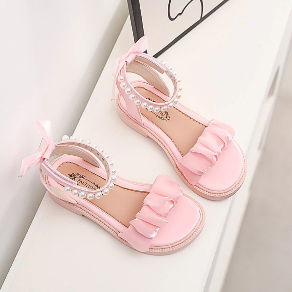 Rosa sandal med volanger för flickor med vita pärlor på en vit möbel och bredvid en vit ram