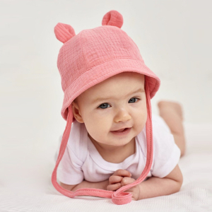 Liten flicka som ligger platt på ett vitt honeycomb-tyg med en vit body och en rosa muslinhatt med öron och snodd.